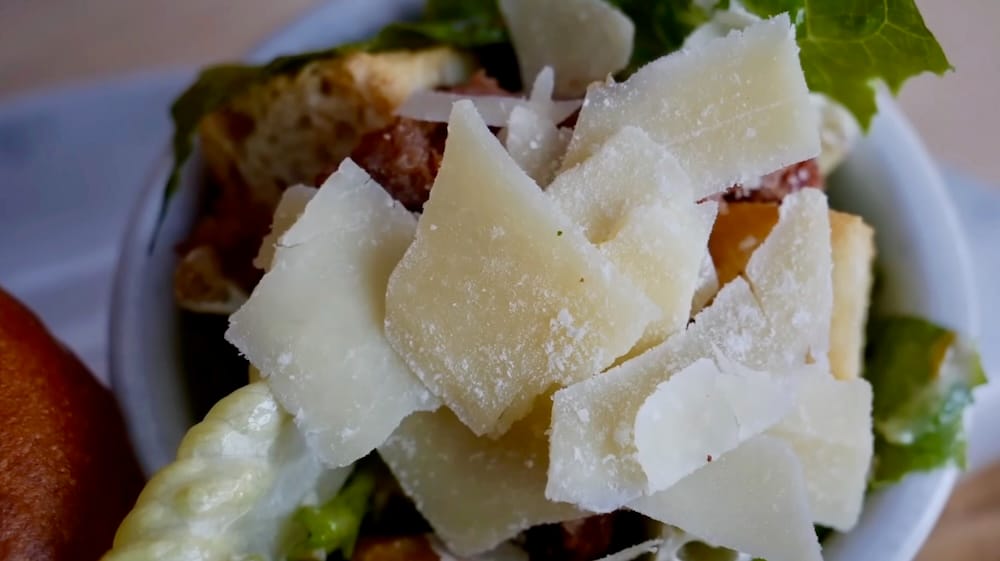Close-up of Caesar salad served at Elk & Oarsman in Banff, Alberta, Canada, topped with large Parmesan shavings, with romaine lettuce and part of the elk burger plate visible around the bowl.