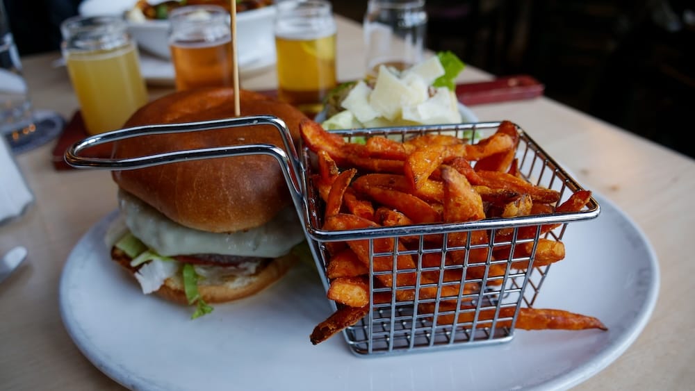 Elk burger meal at Elk & Oarsman in Banff, Alberta, Canada, served with a basket of sweet potato fries and Caesar salad, with part of a shared beer flight visible on the table behind it.