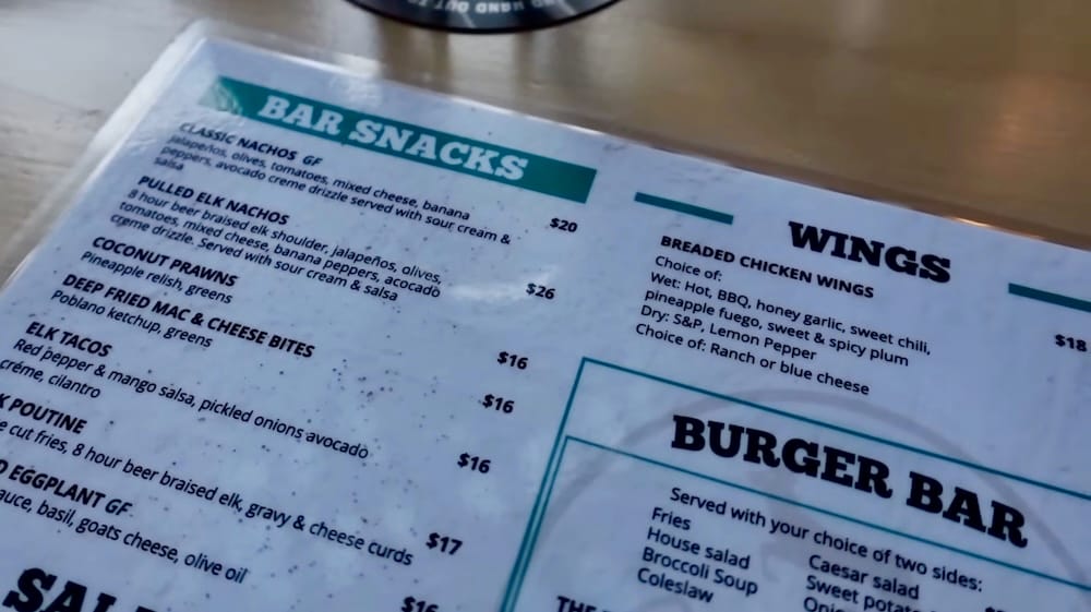 Close-up of the Elk & Oarsman menu in Banff, Alberta, Canada, showing sections for bar snacks, wings and burgers, with menu items including pulled elk nachos, elk tacos and elk poutine.
