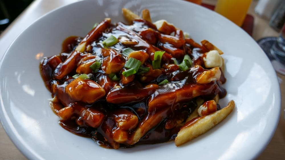 Close-up of poutine at Elk & Oarsman in Banff, Alberta, Canada, with fries covered in dark gravy, cheese curds and chopped green onions in a white bowl, with part of the table setting and a drink softly visible behind it.
