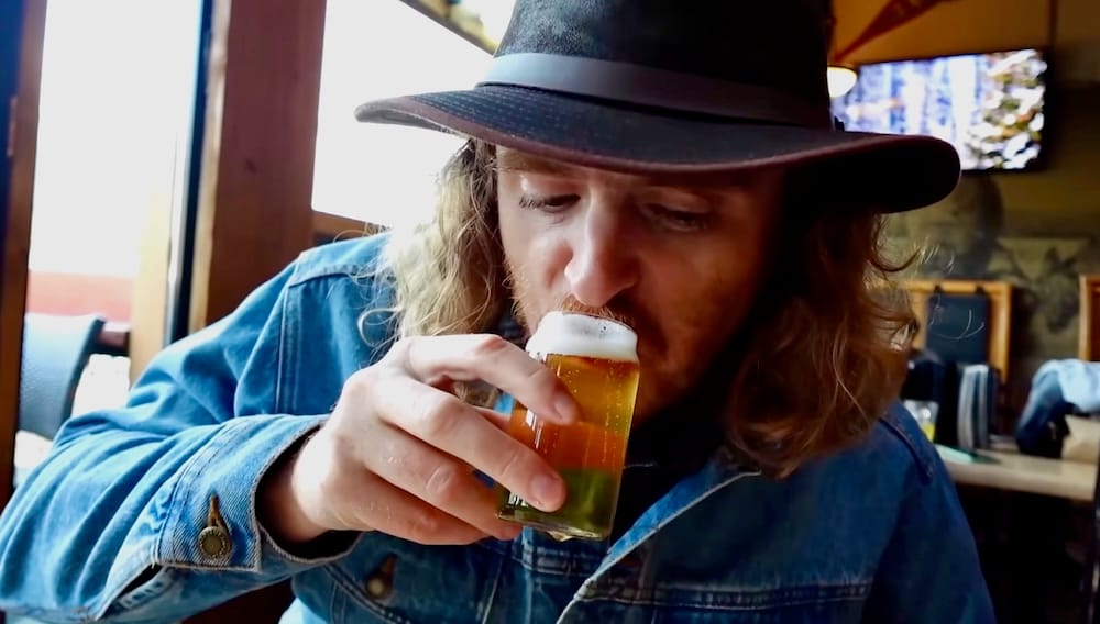 Samuel Jeffery tastes one glass from a beer flight at Elk & Oarsman in Banff, Alberta, Canada, seated beside a window inside the casual sports pub with timber details and dining tables visible behind him.
