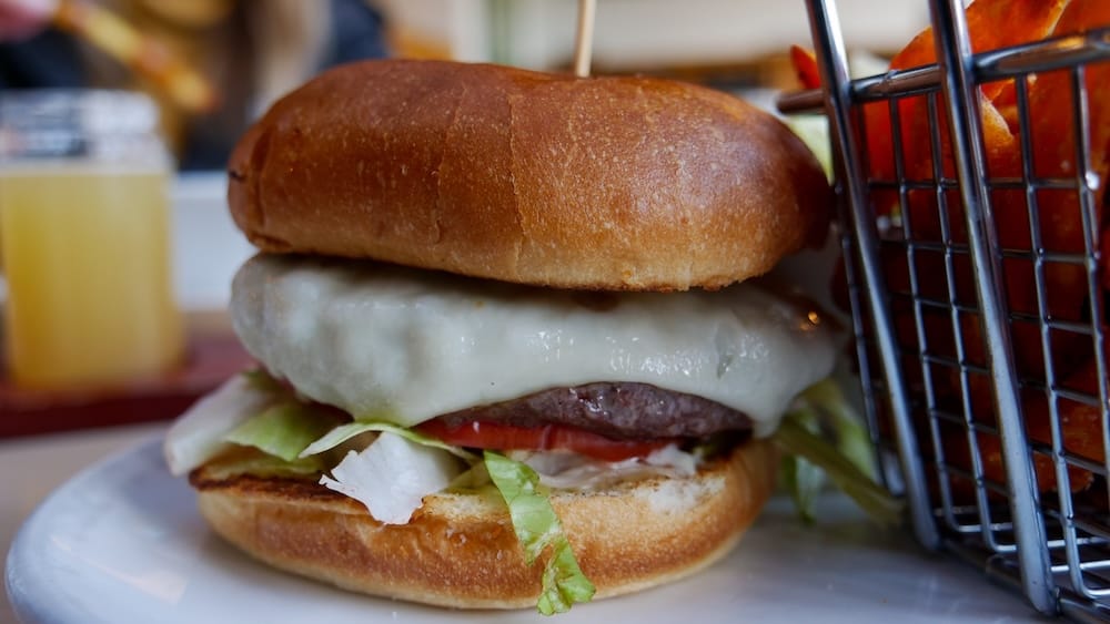 Close-up of an elk burger with melted cheese, lettuce and tomato on a plate at Elk & Oarsman in Banff, Alberta, Canada, with sweet potato fries in a basket and part of a beer flight blurred in the background.