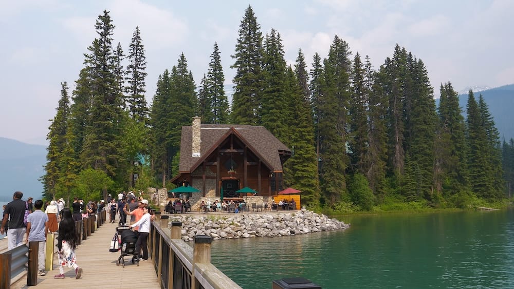 Bridge to Emerald Lake Lodge in Yoho National Park, British Columbia, Canada, beside the lakeshore Pedestrian bridge leading toward Emerald Lake Lodge in Yoho National Park, British Columbia, Canada, with visitors walking beside green lake water, evergreen forest and the lodge ahead.