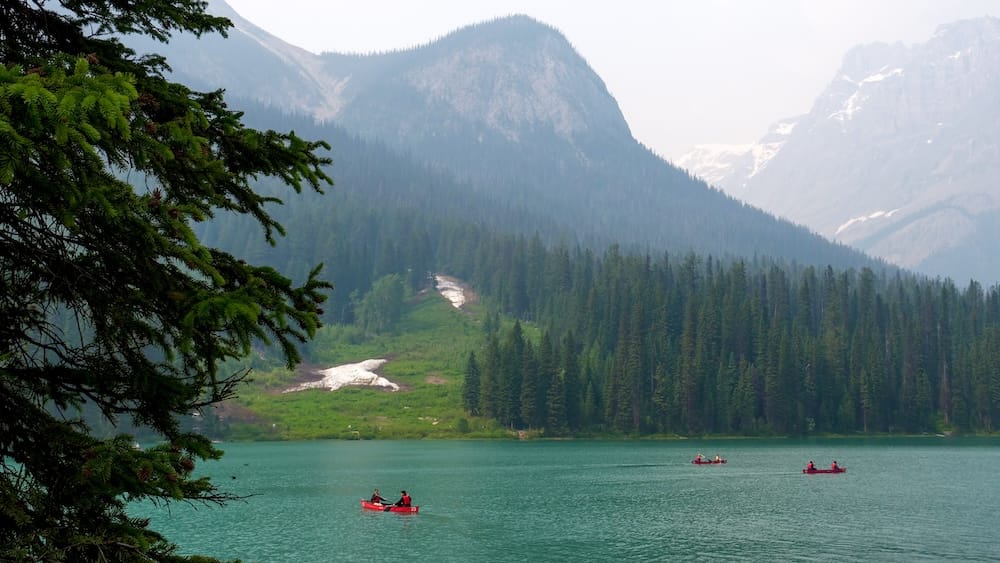 Canoes on Emerald Lake in Yoho National Park, British Columbia, Canada, beneath forested slopes and peaks Several red canoes glide across Emerald Lake in Yoho National Park, British Columbia, Canada, with green water, evergreen forest, lingering snow patches and steep Canadian Rockies mountains in the background.