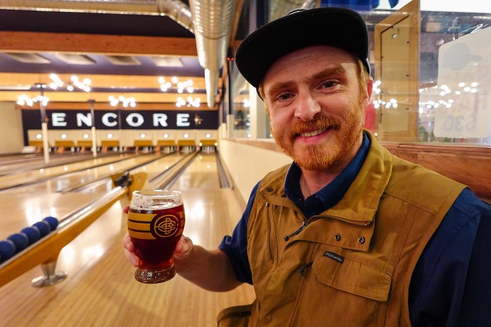 Encore Brewing in Cranbrook, British Columbia, with bowling lanes in the background as Nomadic Samuel enjoys a local craft beer during a relaxed bowling and pizza night, highlighting this brewery as a fun and social stop to include on a Cranbrook day trip.
