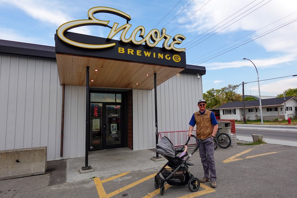 Nomadic Samuel and baby Aurelia at Encore Brewing in Cranbrook Outside Encore Brewing in Cranbrook, BC, Nomadic Samuel stands beside a stroller with baby Aurelia, getting ready for a relaxed family night of bowling, pizza, and craft beer at one of the city’s most welcoming local spots.
