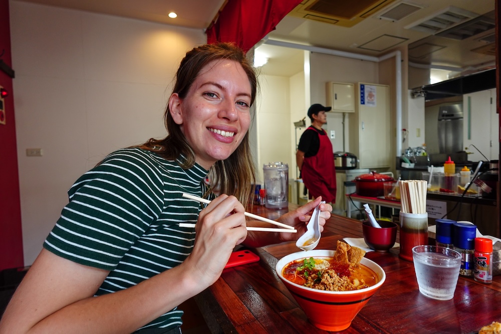 Almuerzo de Enma Ramen picante en una barra de ramen en Noboribetsu Onsen, Hokkaido, Japón – Audrey Bergner Audrey Bergner sonríe con palillos y una cuchara frente a un bol de Enma Ramen picante con caldo rojo y carne picada en la barra de madera de una tienda de ramen en Noboribetsu Onsen, Hokkaido, Japón.