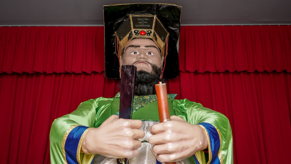 Figura animatrónica del Rey Enma en Enmado, Noboribetsu Onsen, Hokkaido, Japón, con vestimenta ceremonial Primer plano de la figura mecánica del Rey Enma en Enmado, Noboribetsu Onsen, Hokkaido, Japón. Lleva un sombrero ceremonial ornamentado y una túnica verde, y sostiene dos objetos frente a un fondo de cortinas rojas intensas.