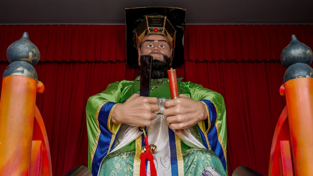 Estatua mecánica del rey Enma con túnica verde dentro del salón Enmado en Noboribetsu Onsen, Hokkaido, Japón La estatua mecánica gigante del rey Enma en Enmado sentada con túnicas verdes y doradas y expresión serena, sosteniendo una tablilla negra y un pergamino rojo frente a una cortina roja en Noboribetsu Onsen, Hokkaido, Japón.