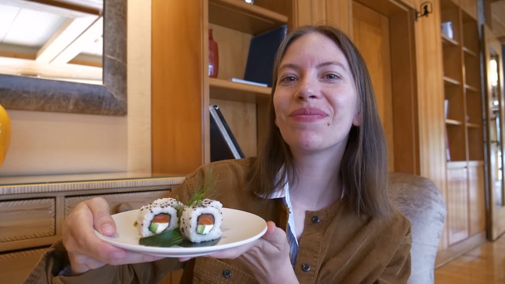 Audrey Bergner Dining on Sushi at Espacio Lago Moreno, Llao Llao Hotel, Bariloche, Río Negro, Patagonia, Argentina Audrey Bergner holds a plate of fresh sushi while dining at Espacio Lago Moreno inside Llao Llao Hotel in Bariloche, Río Negro, Patagonia, Argentina, with warm wood interiors and built-in shelves behind her.
