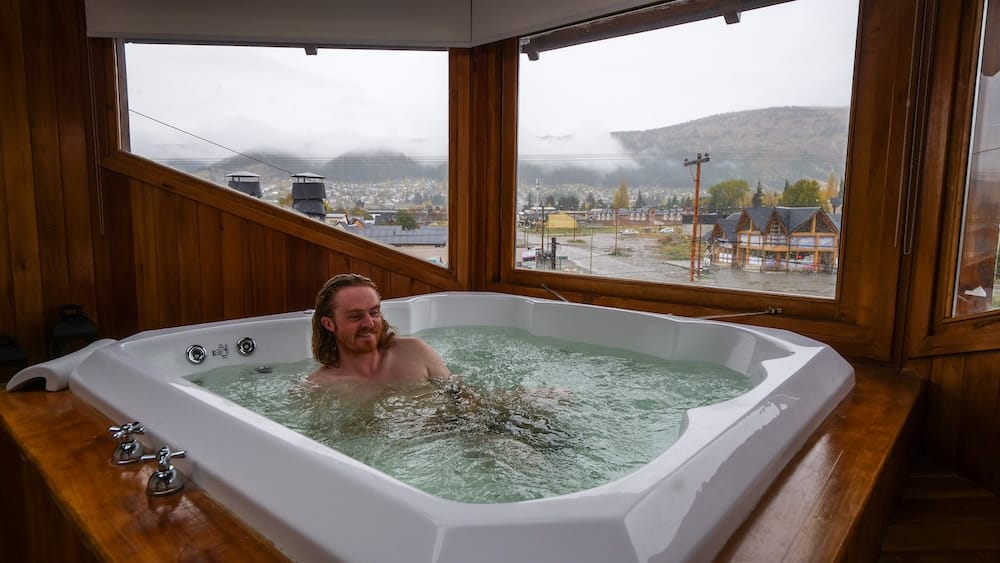 Samuel Jeffery soaks in a jetted hot tub inside a wood-paneled room overlooking rain-soaked rooftops and fog-covered hills in Esquel, Chubut, Argentina, in the Patagonian Andes.