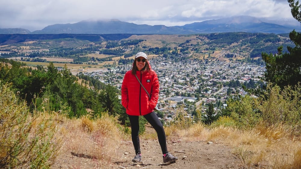 Audrey Bergner wearing a red insulated jacket on a hillside above Esquel, Chubut, Patagonia, Argentina, with the town, dry valley, green slopes and cloud-covered mountains stretching behind her.