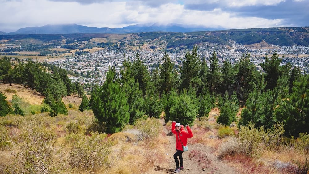 Audrey Bergner Hiking Above Esquel, Chubut, Patagonia, Argentina, Beneath Dramatic Summer Storm Clouds Audrey Bergner bundles up in a red jacket on a hillside trail above Esquel, Chubut, with pine trees, the town below and cloud-covered mountains across Patagonia, Argentina.