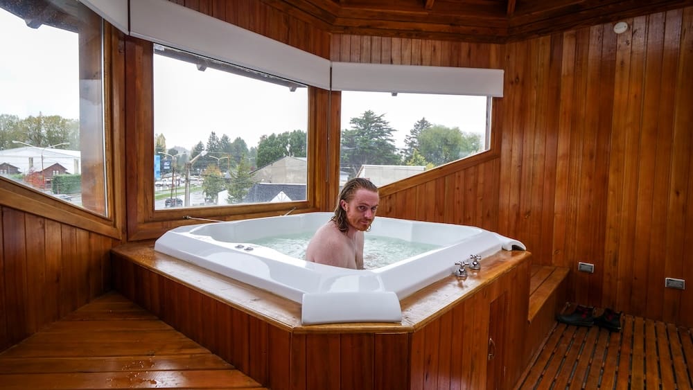 Samuel Jeffery in a Wooden Hot Tub Overlooking Esquel, Chubut, Patagonia, Argentina Samuel Jeffery sits in a wood-paneled corner hot tub overlooking the town of Esquel, Chubut Province, in Argentine Patagonia, with rooftops and trees visible through the windows on an overcast day.
