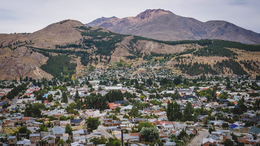 Esquel, Chubut Province, Argentine Patagonia — mountain town panorama beneath forested peaks under overcast sky Aerial panorama of the mountain town of Esquel in Chubut Province, Argentine Patagonia, showing dense rooftops and tree-lined streets beneath a forested ridge and overcast peaks.