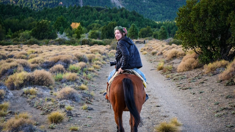Audrey Bergner rides a horse along a forested gravel trail at Estancia Arroyo Verde in Neuquén, Argentine Patagonia, surrounded by scrubland and evergreen trees.