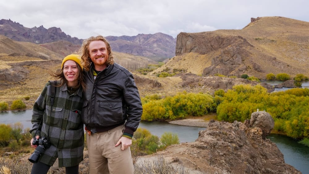 Samuel Jeffery and Audrey Bergner in strong winds at Estancia Arroyo Verde Limay River Patagonia Samuel Jeffery and Audrey Bergner standing in strong Patagonian winds at Estancia Arroyo Verde along the Limay River in autumn, surrounded by rugged hills, winding water, and colorful foliage in Neuquén Argentina.