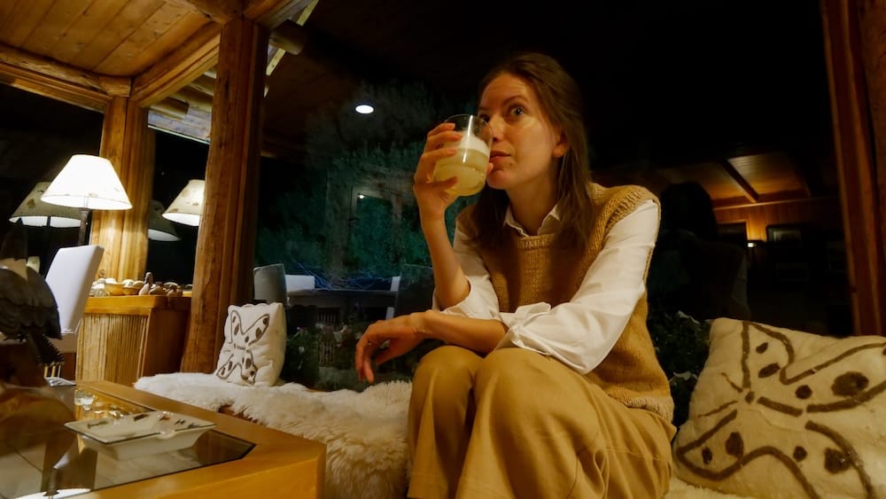 Audrey Bergner sipping a drink inside the warm timber lounge at Estancia Arroyo Verde near Villa Traful, Neuquén, Patagonia, Argentina, with soft lamps, log walls and patterned cushions.