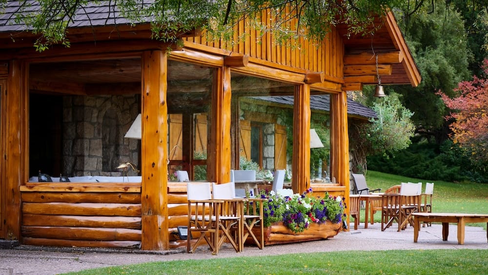 The log-built main lodge at Estancia Arroyo Verde in Neuquén, Argentine Patagonia, with a glass-fronted porch, director's chairs, and a flower-lined terrace overlooking the garden.