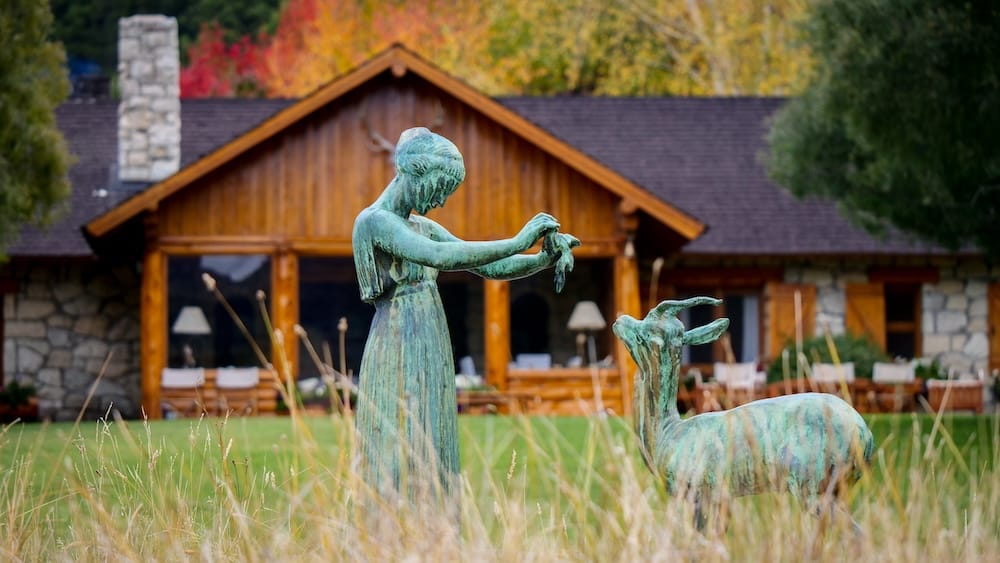 Patinated bronze sculpture of a woman and deer standing in tall grass before the timber-and-stone lodge at Estancia Arroyo Verde near Villa La Angostura, Neuquén, Patagonia, Argentina.