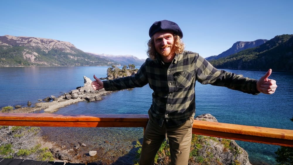 Samuel Jeffery stands at a wooden viewpoint above a clear Andean lake at Estancia Arroyo Verde in Neuquén, Patagonia, Argentina, with rocky shoreline, forested slopes and broad mountain ridges behind him.