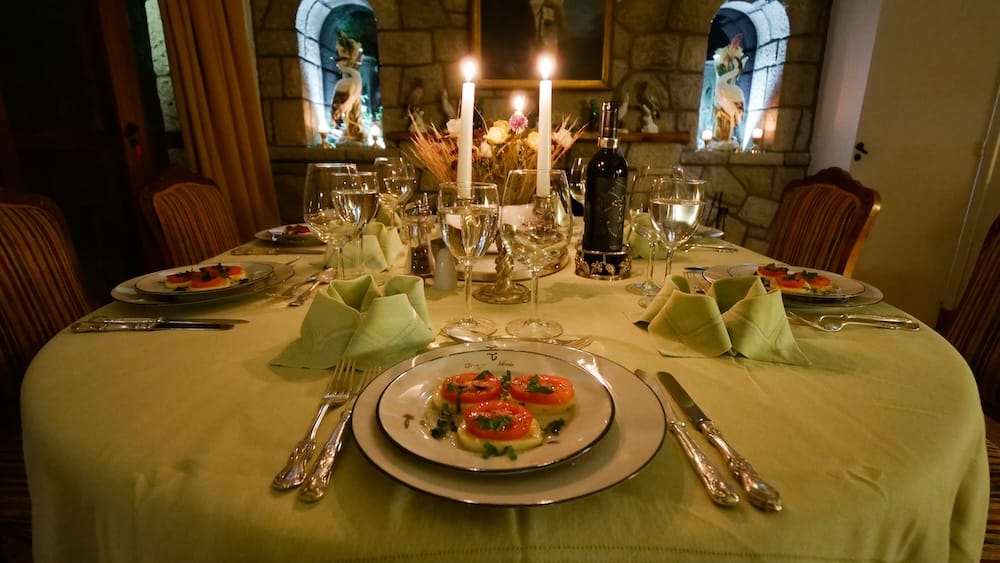 A candlelit dining table is set inside Estancia Arroyo Verde in Neuquén, Patagonia, Argentina, with wine glasses, plated starters, a bottle of wine and rustic stone walls framing the warm lodge interior.