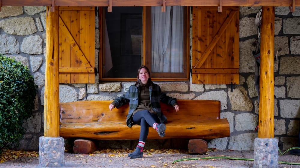 Audrey Bergner sits on a carved wooden bench outside Estancia Arroyo Verde in Neuquén, Patagonia, Argentina, framed by rustic stone walls, timber posts, wooden shutters and fallen autumn leaves.