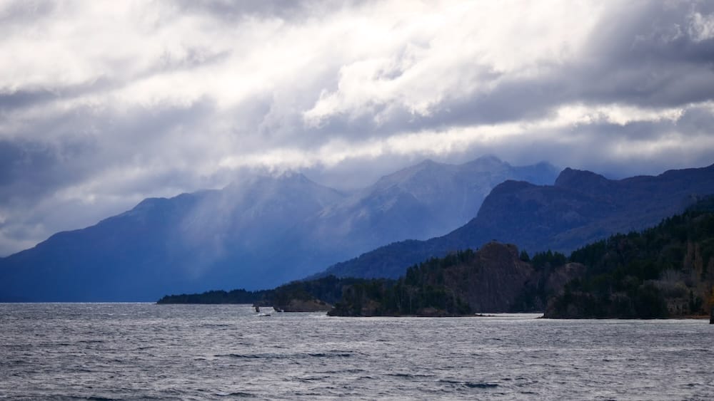 Storm Clouds Over Estancia Arroyo Verde, Neuquén, Patagonia, Argentina Dramatic storm clouds sweep over the lake and mountain peaks at Estancia Arroyo Verde in Neuquén, Patagonia, Argentina, with dark water and forested shoreline beneath shafts of light.