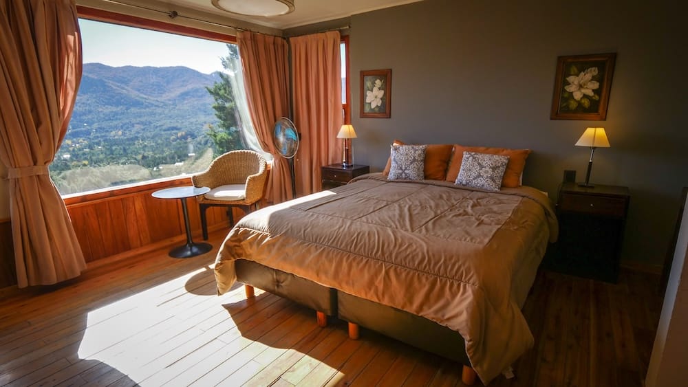 A sunlit bedroom at Estancia Del Carmen near Bariloche, Río Negro, Argentine Patagonia, with a large window framing forested mountains above the Lago Gutiérrez valley.