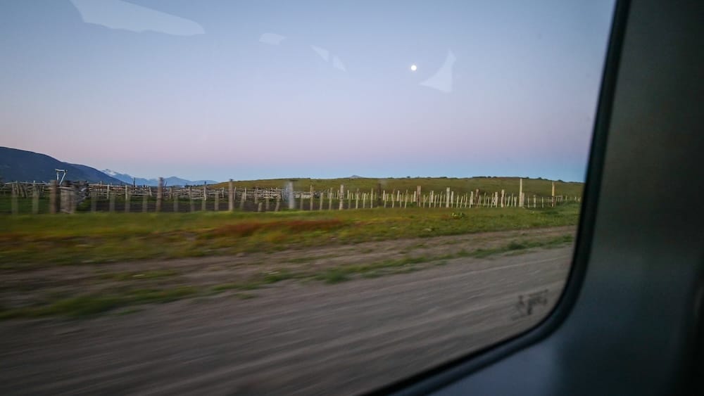 Rustic fenced pastures at Estancia Nibepo Aike near El Calafate, Santa Cruz, Patagonia, Argentina, seen through a vehicle window beneath a fading moon and pale evening sky.