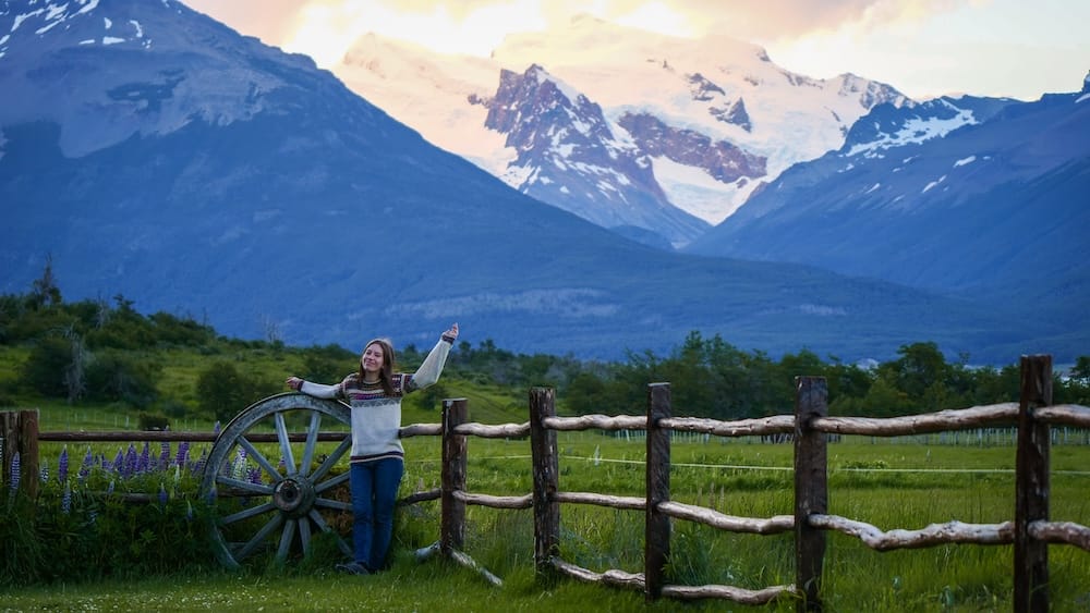 Audrey Bergner leans on an old wagon wheel beside a wooden fence at Estancia Nibepo Aike in Santa Cruz, Argentine Patagonia, with lupine wildflowers and snow-covered glacial peaks behind her.