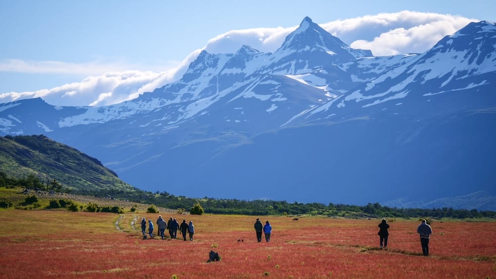 A small group walks across red grasslands at Estancia Nibepo Aike in Santa Cruz, Patagonia, Argentina, with a dramatic snow-covered mountain range rising beyond the open ranch landscape.