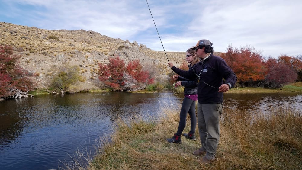 Audrey Bergner stands beside a fishing guide on the grassy riverbank at Estancia Tecka in Chubut, Patagonia, Argentina, learning fly casting beside calm water with autumn shrubs and dry hills in the background.