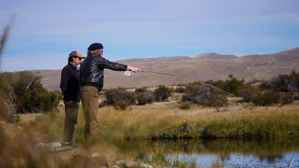 Samuel Jeffery practises fly casting beside a guide on the riverbank at Estancia Tecka in Chubut, Patagonia, Argentina, with still water, golden grass, low shrubs and open steppe hills stretching across the background.