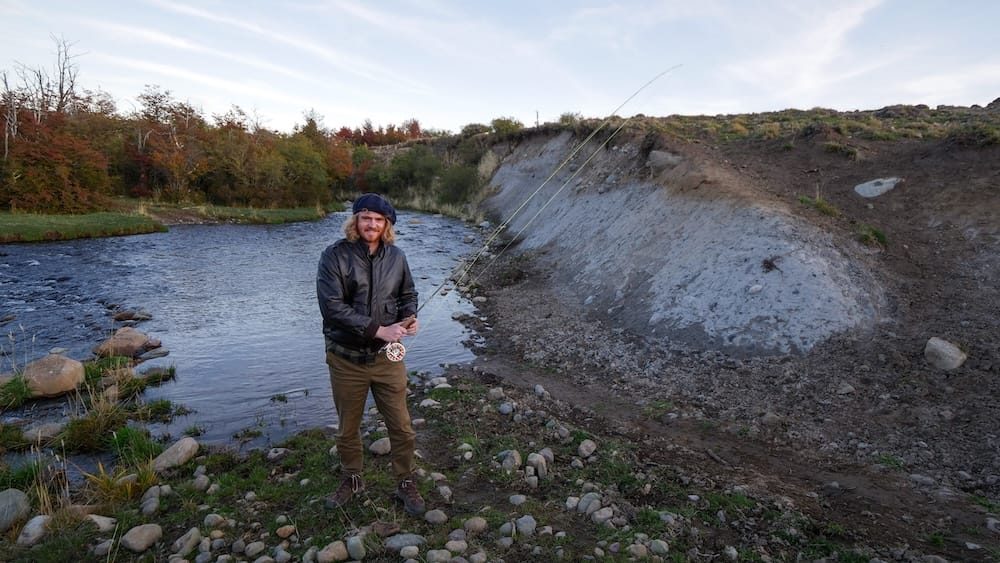Samuel Jeffery stands beside a winding river at Estancia Tecka in Chubut, Patagonia, Argentina, holding a fly-fishing rod with rocky banks, autumn foliage and open ranch terrain surrounding him.