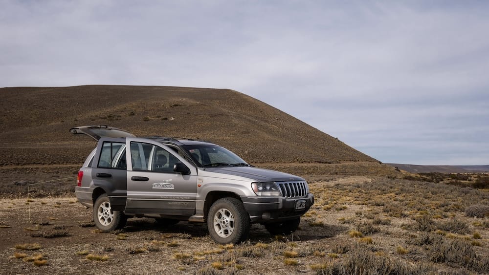A silver Jeep sits on the open steppe at Estancia Tecka in Chubut, Patagonia, Argentina, surrounded by dry grassland, low rolling hills and a broad overcast sky in a remote ranch landscape.