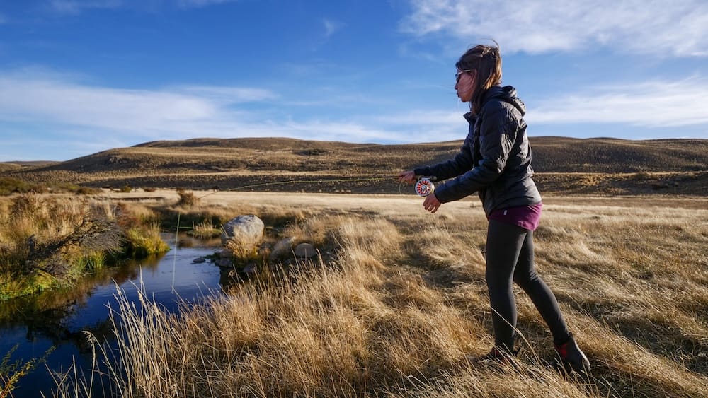 Audrey Bergner practises fly fishing beside a narrow stream at Estancia Tecka in Chubut, Patagonia, Argentina, with dry grassland and rolling steppe beneath a wide blue sky.