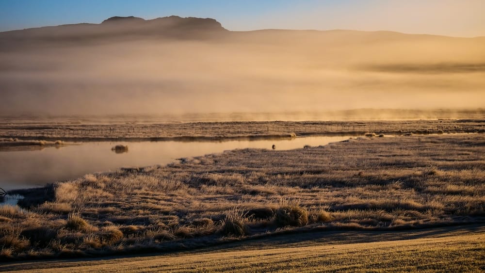 Golden morning mist rolls over frost-covered grassland and a still river channel at Estancia Tecka near Esquel, Chubut, Argentine Patagonia, with low mountains silhouetted in the distance.