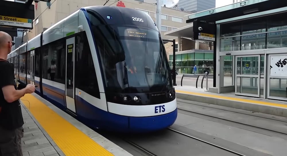 Un tren del tren ligero (LRT) ETS Valley Line llega al andén de la calle 100 Street en el centro de Edmonton, Alberta, con un pasajero filmando desde el borde del andén y el horizonte de la ciudad de fondo.