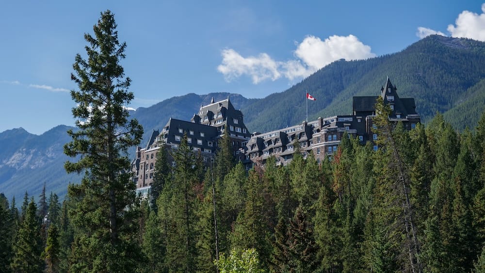 Fairmont Banff Springs rising above dense evergreen forest in Banff, Alberta, Canada, with layered Canadian Rockies mountains forming the backdrop beneath a clear blue sky.