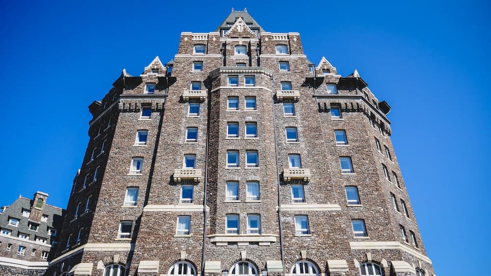 A close upward view of the Fairmont Banff Springs Hotel in Banff, Alberta, Canada, showing its tall stone tower, rows of windows and château-style architecture against a clear blue sky.