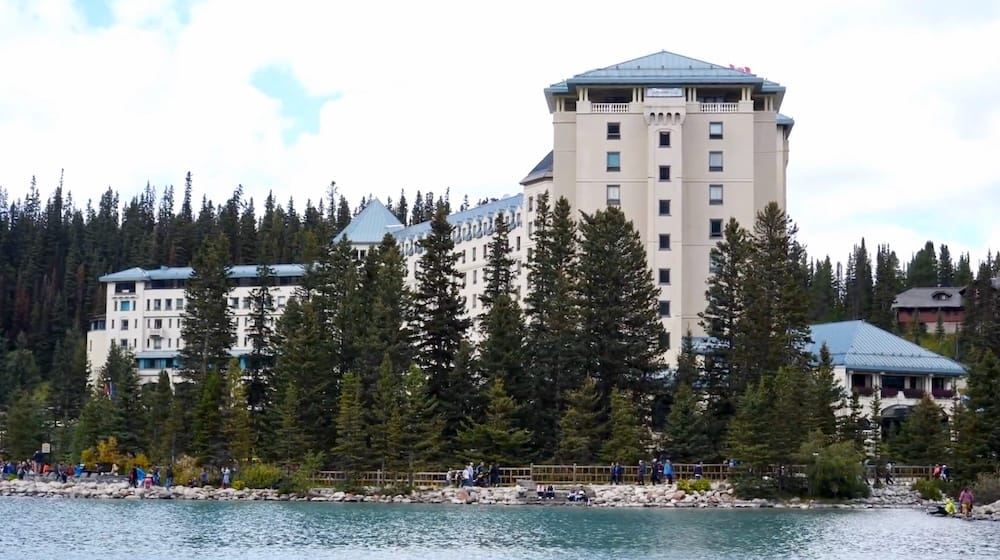 Fairmont Chateau Lake Louise stands behind evergreen trees on the shore of Lake Louise in Banff National Park, Alberta, Canada, with visitors gathered along the lakeside path.