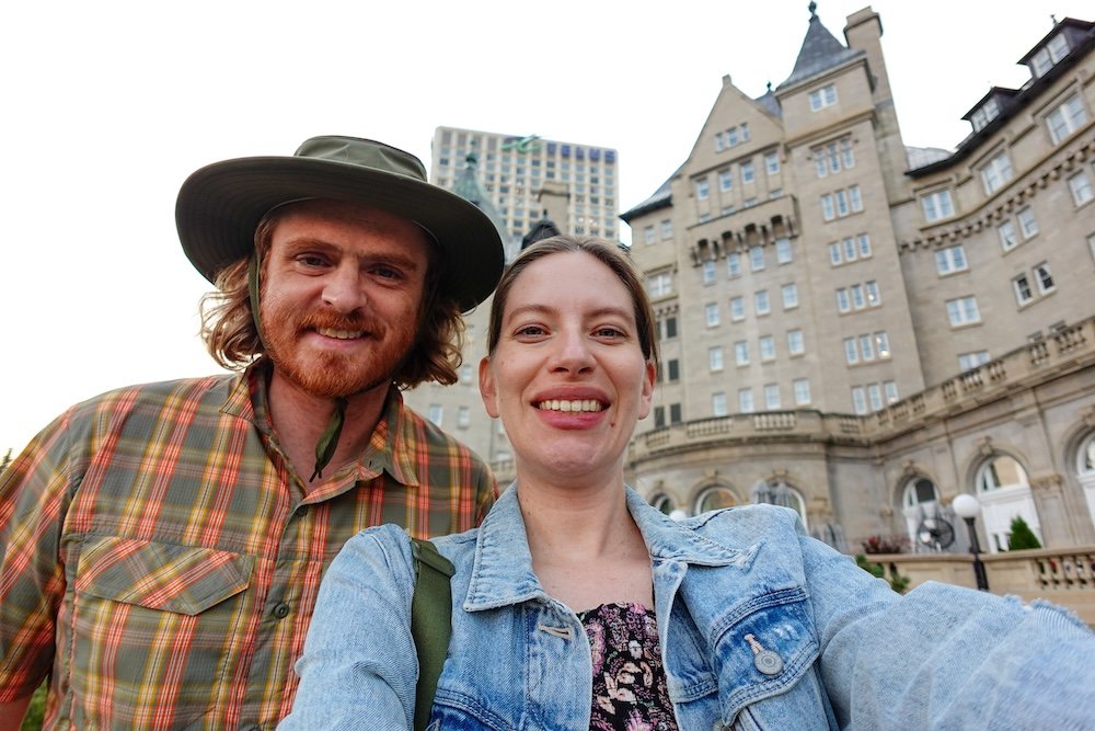 Nomadic Samuel and Audrey at Fairmont Hotel Macdonald in Edmonton Audrey Bergner of That Backpacker and Nomadic Samuel Jeffery smile in front of the historic Fairmont Hotel Macdonald in Edmonton during their late-summer babymoon, capturing a relaxed moment on the castle’s riverside terrace before an evening stroll.