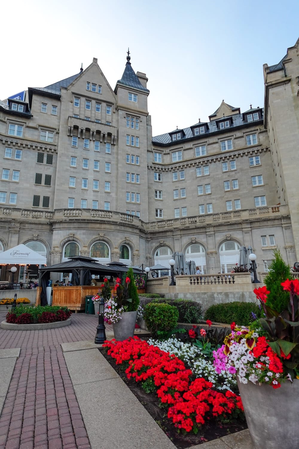 Fairmont Hotel Macdonald Exterior and Garden Terrace, Downtown Edmonton, Alberta, Canada The chateau-style stone exterior of the Fairmont Hotel Macdonald in downtown Edmonton, Alberta, Canada, with turrets, a terrace, and flowerbeds of red and white blooms in the foreground.