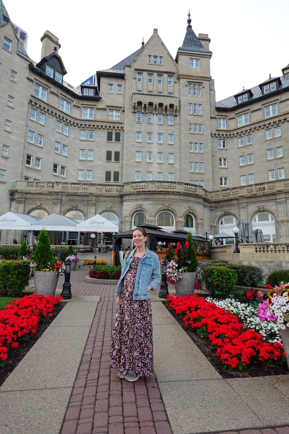 Audrey Bergner parada en el camino de ladrillos que atraviesa el jardín florecido del patio del Fairmont Hotel Macdonald en el centro de Edmonton, Alberta, Canadá, con canteros de flores rojas y blancas y la fachada de piedra estilo castillo del hotel de fondo.