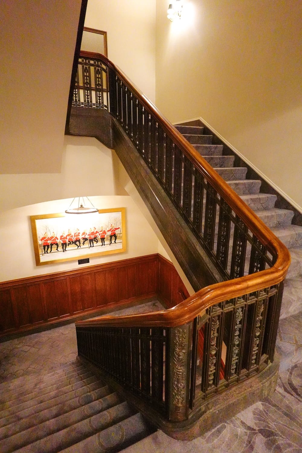 Gran escalera histórica y paneles de madera interiores en el Fairmont Hotel Macdonald en Edmonton, Alberta, Canadá Una vista interior mirando hacia abajo por una gran escalera alfombrada con un pasamanos de madera oscura ornamentado, balaustres de hierro forjado y obras de arte enmarcadas en el descanso dentro del Fairmont Hotel Macdonald en Edmonton, Alberta, Canadá.