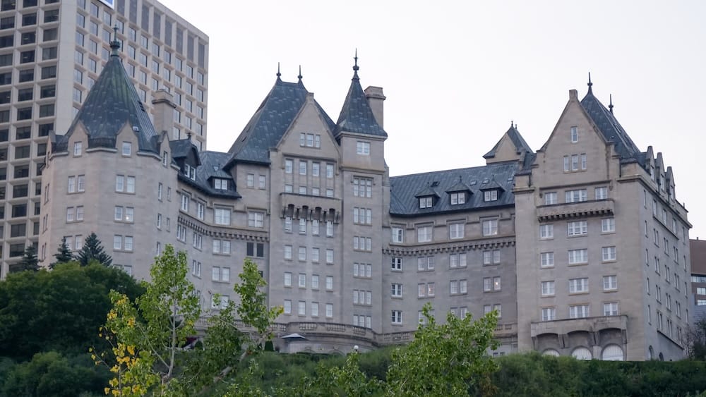 Exterior tipo château del Fairmont Hotel Macdonald desde el valle del río North Saskatchewan en Edmonton, Alberta, Canadá La gran fachada de piedra caliza y las torretas de cobre del Fairmont Hotel Macdonald se elevan sobre los árboles verdes del valle del río con un rascacielos moderno de fondo en Edmonton, Alberta, Canadá.