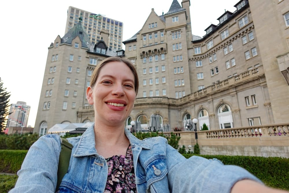 Audrey Bergner smiling for a selfie outside Fairmont Hotel Macdonald in Edmonton, Alberta, Canada, with the château-style stone façade, terraces and downtown towers visible behind her.