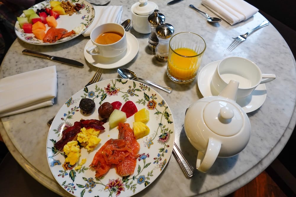 Desayuno del Fairmont Gold Lounge en el Fairmont Hotel Macdonald, Edmonton, Alberta, Canadá, con salmón ahumado y fruta fresca Un desayuno servido en el Fairmont Hotel Macdonald en Edmonton, Alberta, Canadá, sobre una mesa de mármol, con salmón ahumado, huevos revueltos, panceta, fruta fresca, una tetera y taza blancas, jugo de naranja y servilletas de tela dobladas.