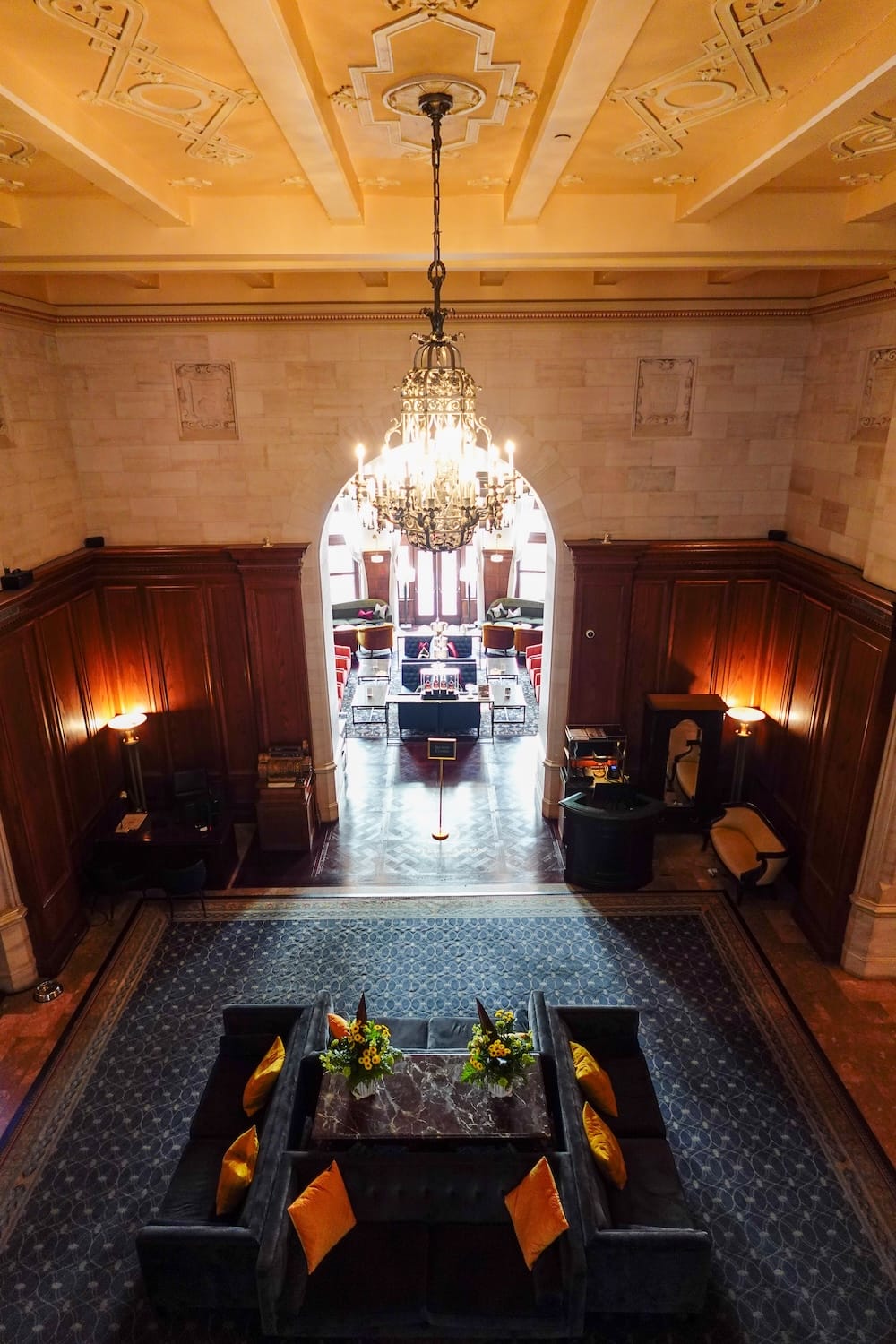 Vista del balcón del lobby lounge en el Fairmont Hotel Macdonald, Edmonton, Alberta, Canadá, con candelabro de hierro forjado y piso en espiga Una vista elevada desde el balcón hacia un lounge en el Fairmont Hotel Macdonald en Edmonton, Alberta, Canadá, mostrando un techo de yeso ornamentado, un gran candelabro de hierro forjado, un piso de madera en espiga, paneles de madera oscura y una sala de estar con sofás azules y almohadones naranjas.